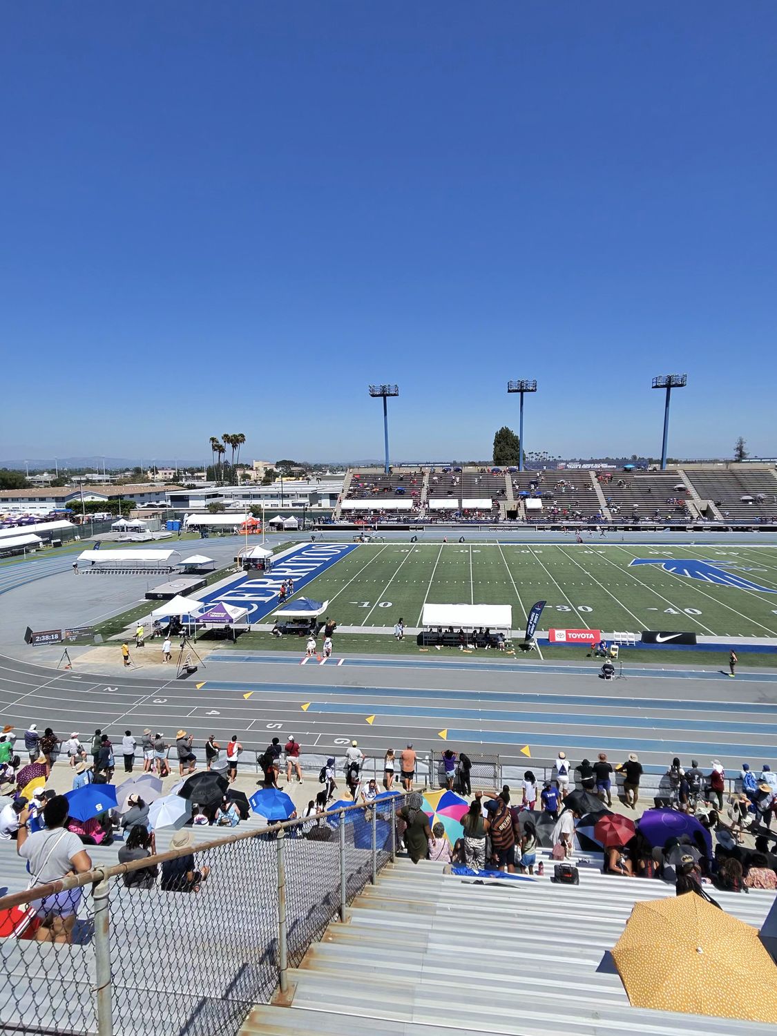 Wide view of the track and field stadium during competition with spectators in the stands and athletes on the blue track