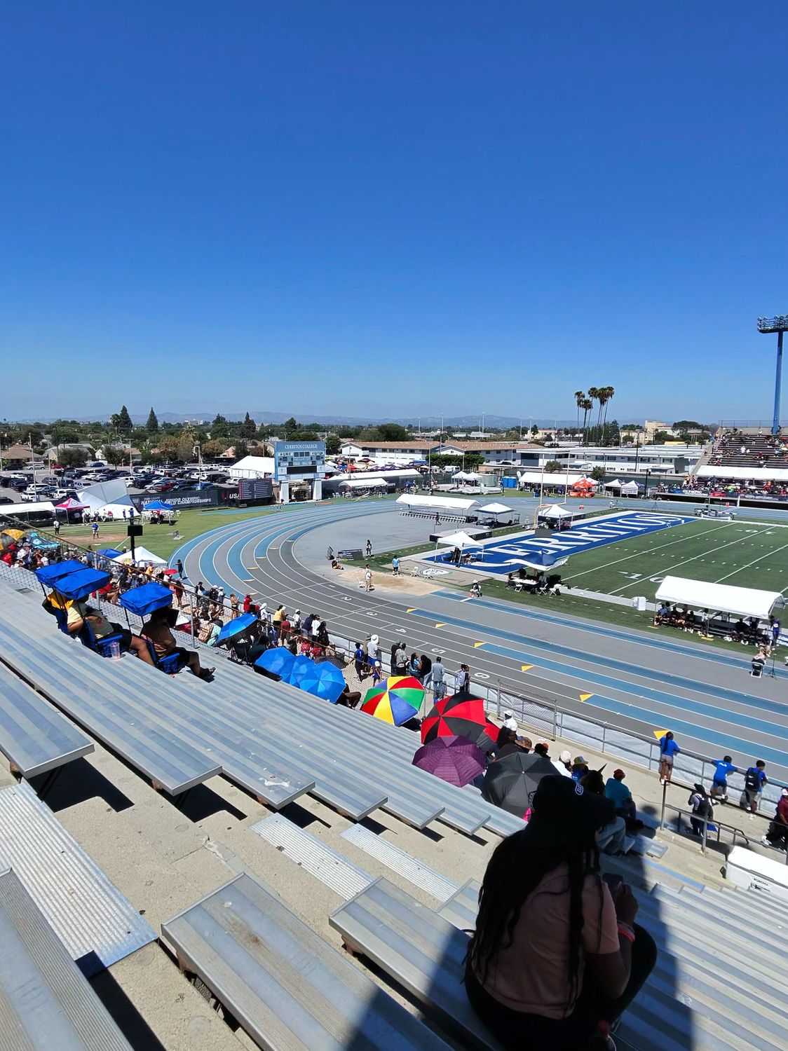 Spectators in bleachers with portable stadium seats that have sunshade canopies providing shade at a track meet