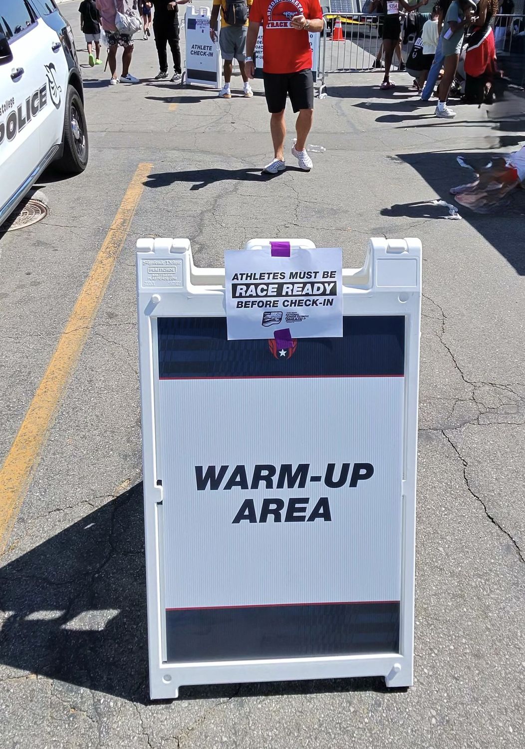 Warm-up area entry point sign reading Athletes Must Be Race Ready Before Check-In with police vehicle nearby